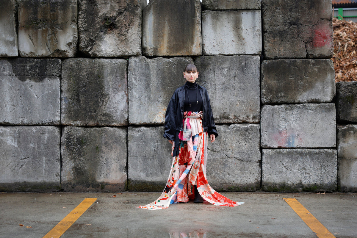 By a concrete wall: a skirt of many layered kimono fabrics, its hem trailing long.