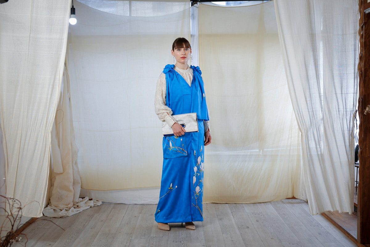 In a room with white curtains, she carries a clutch with a blue dress painted with white flowering branches.