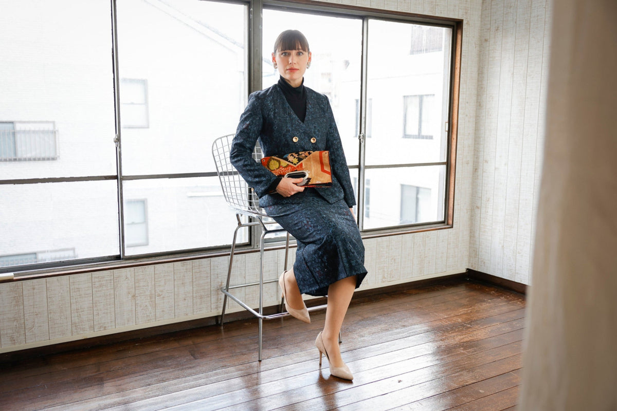 On a wire-backed high stool by the window, she holds an obi clutch at her waist, hand through the handle.