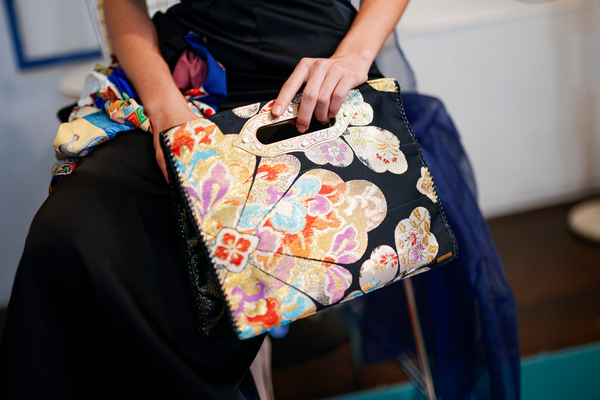 The MIKOSHI Bag rests on her knees, fingertips hooked over the handle.