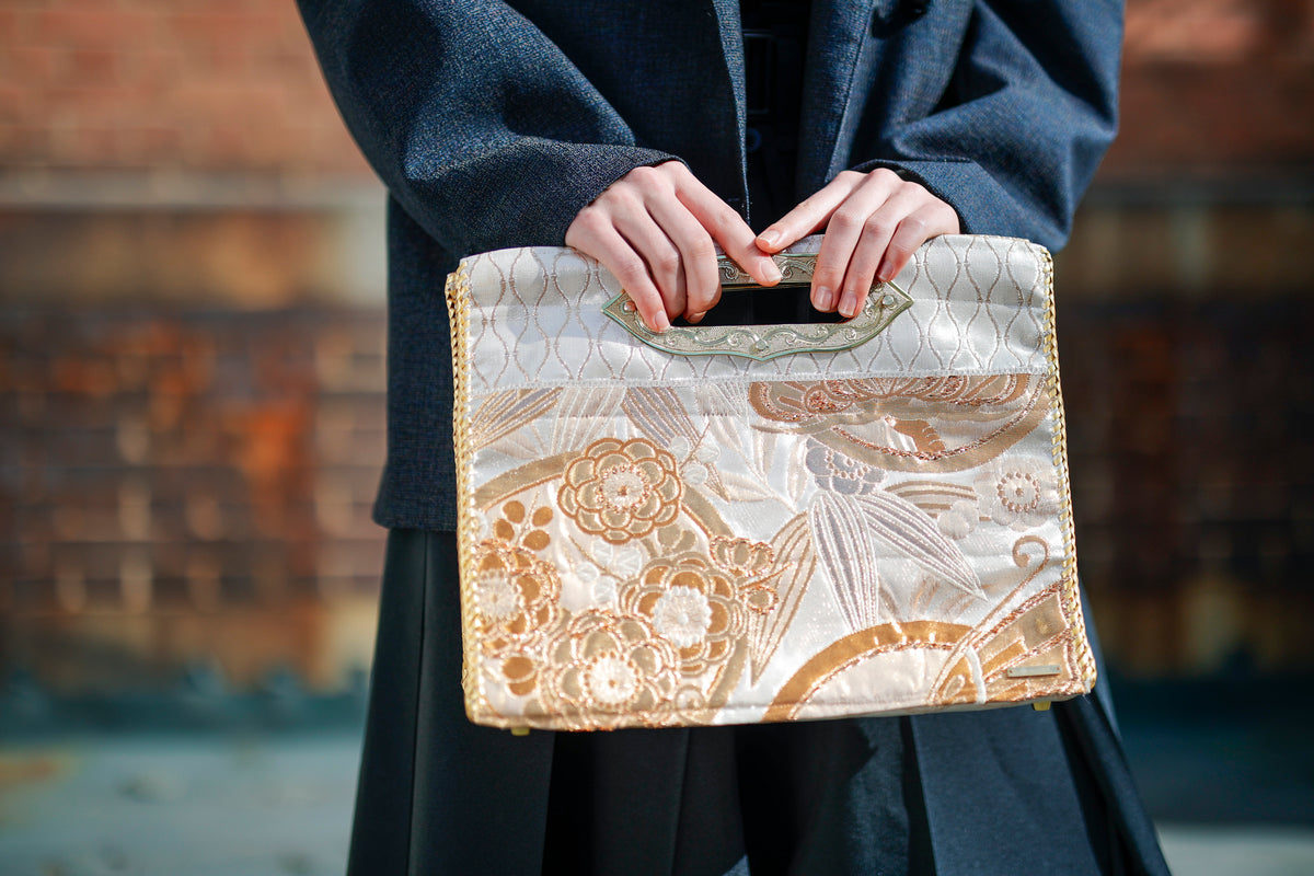 Two hands grip the handle. Wavy lines run through the ground weave above, with gold flowers below.