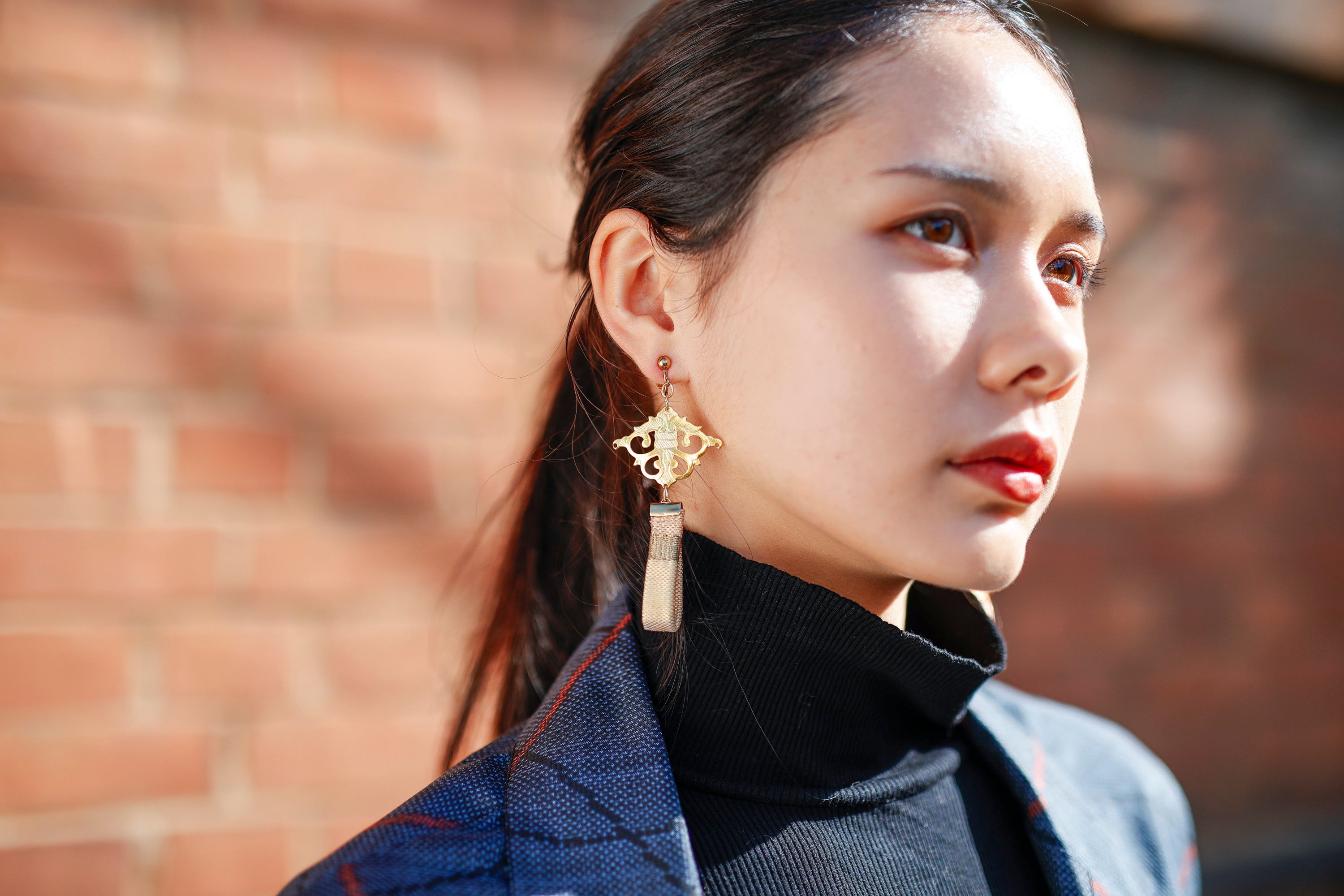 Hana-warabe earring with a flat natural-coloured tassel, against a red brick wall