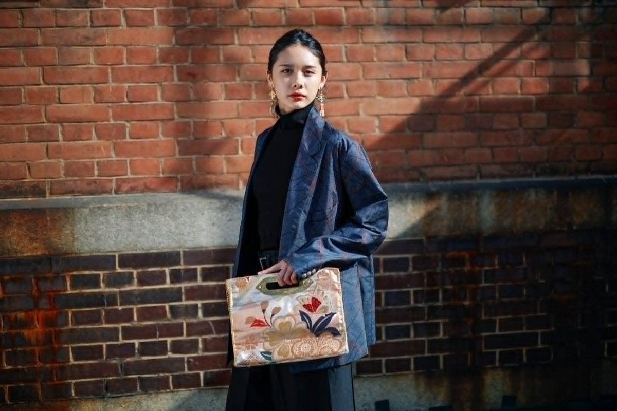 A model holding the MIKOSHI Series bag in front of a brick wall